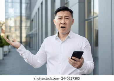 Close-up portrait of an angry young Asian man in a white shirt standing outside an office building, holding a phone and throwing his hands up in frustration and looking at the camera. - Powered by Shutterstock