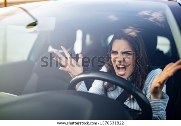 Closeup\
portrait, angry young sitting woman pissed off by drivers in front\
of her and gesturing with hands. Road rage traffic jam concept.\
Woman is driving her car very\
aggressive
