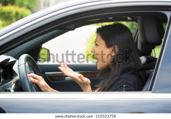 Closeup portrait, angry\
young sitting woman pissed off by drivers in front of her and\
gesturing with hands, isolated city street background. Road rage\
traffic jam concept.