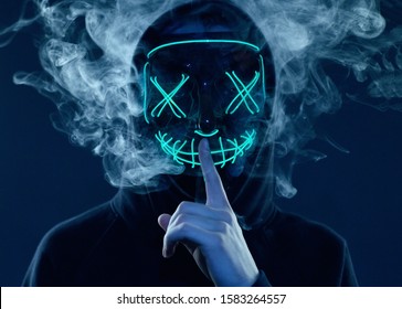 Close-up portrait of an anonymous man hiding his face behind a scary neon mask in a colored smoke. Studio shot - Powered by Shutterstock