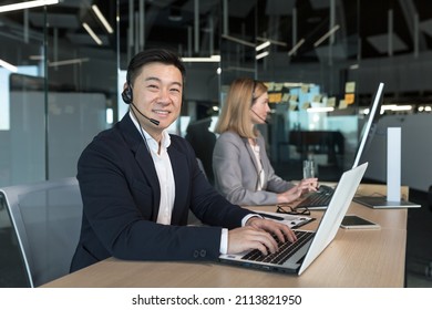 Close-up portrait of Asian man in business attire, call center employee looks at camera and smiles, businessman uses computer for video call and headset