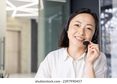 Close-up portrait of Asian online support worker, woman smiling and looking at camera using headset phone for online consultation and video call, working inside office with laptop. - Powered by Shutterstock