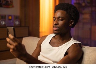 Close-up Portrait Of Athletic, Muscular Man Relaxing On A Couch In Sunset-lit Living Room, Boy With A Serious Expression Is Talking On The Phone To A Friend On A Video Camera