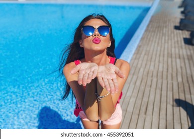 Close-up portrait of attractive brunette girl with long hair standing near pool. She wears pink T-shirt, sunglasses.  She stretches her hands to the camera and makes a kiss.