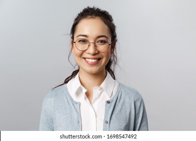 Close-up portrait of attractive, friendly-looking asian female office worker, employee or teacher in glasses, smiling broadly camera with enthusiastic attitude, stand grey background