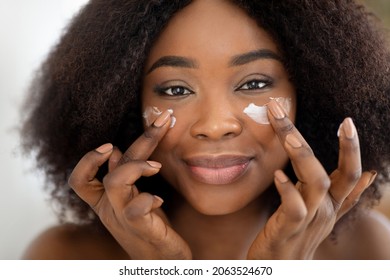 Closeup Portrait Of Attractive Millennial Black Lady Applying Facial Cream Under Her Eyes. Pretty African American Woman Taking Care Of Skin, Putting On Nourishing Lotion. Beauty And Natural Cosmetics