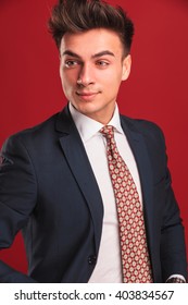 Closeup Portrait Of Attractive Young Businessman In Black Suit With Red Tie, With Open Jacket, Posing In Red Studio Background While Looking Away From The Camera