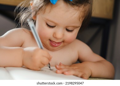 Close-up Portrait Of A Baby Girl Drawing With Pencil Sitting At A Wooden Table. Baby Artist Activity Lifestyle Concept. Baby Development.