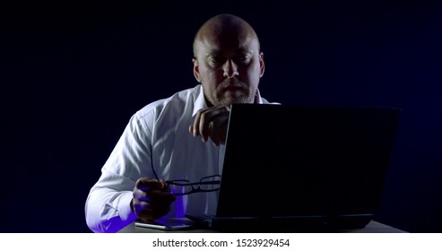 Close-up Portrait Of A Bald Man In Glasses And A Shirt, He Is Sitting At A Laptop, Emotions, Happy Looking At The Computer Screen, Rooting For Someone, Next To Him On The Table Is A Mobile Phone.