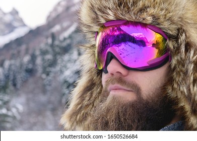 Close-up portrait of a bearded happy snowboarder skier in a ski mask with goggles and a fur big old-school hat on a background of a winter snowy mountains