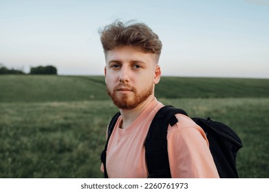 Close-up portrait of a bearded young man in casual clothes and with a backpack standing in a field overgrown with grass and looking at the camera with a serious face - Powered by Shutterstock