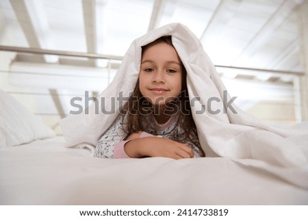 Close-up portrait of a beautiful Caucasian little child girl 6 years old, lying on the bed under white blanket duvet while waking up in the morning, smiling cutely looking at the camera.