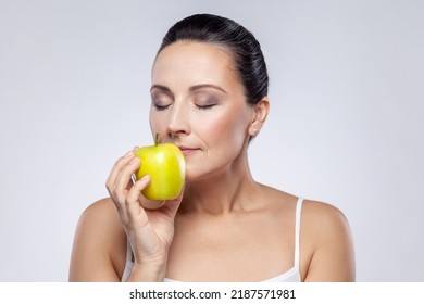 Closeup Portrait Of Beautiful Charming Middle Aged Woman Holding Smelling Green Apple, Vitamins For Beauty And Skin Care, Keeps Eyes Closed. Indoor Studio Shot Isolated On Gray Background.