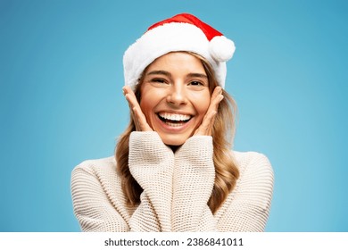 Closeup portrait of beautiful cute happy woman wearing Santa hat holding hands on cheeks looking at camera isolated on blue background.  Christmas, holidays celebration concept - Powered by Shutterstock