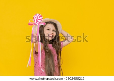 A close-up portrait of a beautiful fashionable cheerful little girl in a straw hat and pink dress, eating a sweet candy lollipop on a yellow background. On an isolated background.