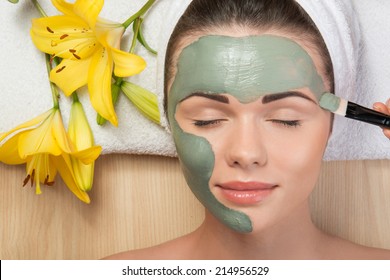 Close-up portrait of beautiful girl with closed eyes  with a towel on her head applying facial clay mask and beauty treatments lying on a table in spa near yellow flower