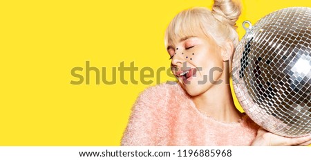 Close-up portrait of beautiful girl fooling around with silver ball. Blonde model with sparkles on face. Festive party concept. Isolated on yellow background