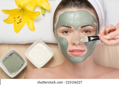 Close-up portrait of beautiful girl looking at the camera with a towel on her head applying facial clay mask and beauty treatments lying on a table in spa near yellow flower and two plates