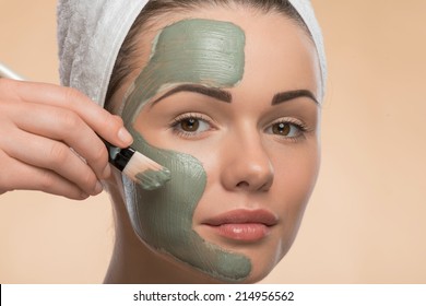 Close-up Portrait of beautiful looking at the camera girl in spa with a  towel on her head applying facial clay mask and beauty treatments with brush on her face isolated on beige background