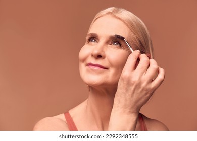 Close-up portrait of beautiful senior woman holding eyebrow makeup stick, smiling on beige background and looking away. Skin care and makeup concept. - Powered by Shutterstock