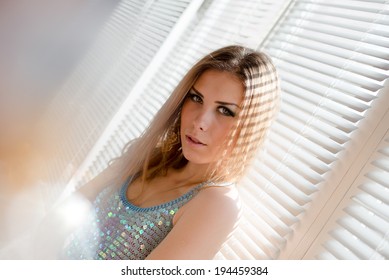 Closeup Portrait Of Beautiful Sensual Blond Young Woman With Green Eyes Relaxing Looking Into The Camera On Sun Shining Flare Through Window Blinds Background