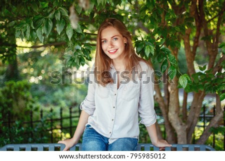 Closeup portrait of beautiful smiling white Caucasian girl woman with long blonde hair and blue eyes wearing white shirt and jeans outside in summer park in green foliage trees looking in camera.