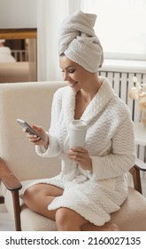 Closeup Portrait Of Beautiful And Smiling Young Woman In A White Robe And With A Towel After Spa On Her Head Using Mobile Phone For Social Media, Drinking Takeaway Coffee In Hotel Room