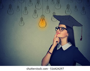 Closeup portrait beautiful thoughtful graduate student girl young woman in cap gown looking up at bright light bulb thinking isolated gray wall background. Graduation ceremony future career concept - Powered by Shutterstock