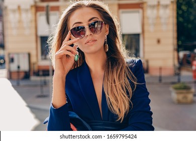 close-up portrait of beautiful woman dressed in stylish blue jacket walking in autumn sunny street wearing elegant sunglasses