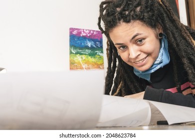 Close-up Portrait Of Beautiful Young White Girl Hispanic Latina Art Director With Dreadlocks, Smiling, In Her Atelier, Leaning On Her Desk With Papers And A Picture Of The Gay Pride Flag On The Wall.