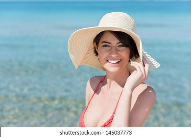 Closeup portrait of a a beautiful young woman posing on a beach in a swimsuit and wide brimmed floppy straw sunhat as she protects herself gainst the UV rays of the sun