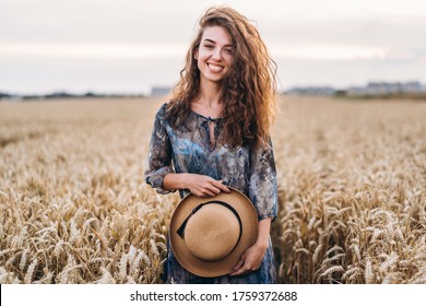 Closeup portrait of a beautiful young woman with curly hair. Woman in dress and hat standing in wheat field. - Powered by Shutterstock