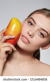 Close-up Portrait Of A Blond Girl With Naked Shoulders. She Holding Mango Near Her Cheek. Girl Has Nude Makeup With Black Arrows. The Photo Is Taking On Grey Background. 