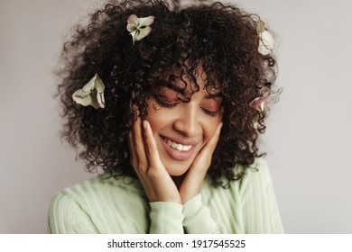 Close-up portrait of brunette curly woman with flowers in hair smiling and looking down. Pretty dark-skinned lady poses on white background.