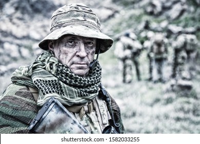 Close-up portrait of brutal commando veteran, experienced army commander or officer with dirty face, wearing camouflage bonnie, shemagh, tactical radio headset with microphone, looking in camera