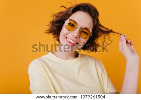 Close-up portrait of carefree white girl plays with short wavy hair. Photo of elegant european woman with black tattoo isolated on yellow background.