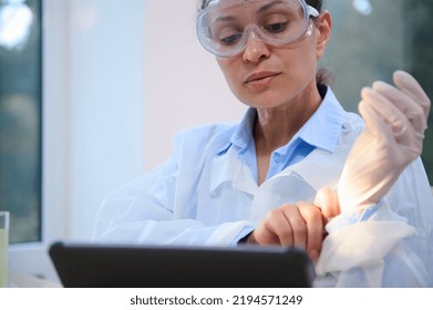 Close-up Portrait Of A Charming Multi-ethnic Middle-aged Woman, Scientist, Chemist, Medical Laboratory Worker In White Lab Coat And Safety Goggles Works On Digital Tablet And Puts On Protective