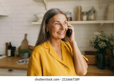 Close-up Portrait Of Charming Retired Lady In Yellow Shirt Having Nice Phone Conversation Isolated Over Kitchen Interior Background, Looking Through Window During Talk On Black Smartphone