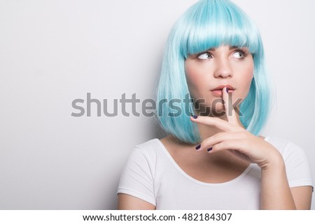 Closeup portrait of cheeky young girl in modern futuristic style wearing blue wig with curious expression and finger on lip while looking sideways over white wall background with copyspace