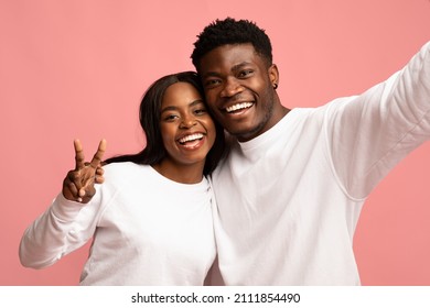 Closeup portrait of cheerful beautiful millennial black couple in white hugging and smiling, taking selfie together on pink studio background. Love, affectionate, family, relationships concept