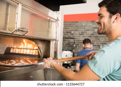 Close-up Portrait A Cheerful Chef Wearing Gloves Putting Raw Prepared Pizza In Oven Using Peel In Pizza Shop