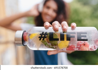 Close-up portrait of cheerful girl in headphones with natural juice with fruits and smiling. Blur photo of pretty young woman with elegant manicure holding bottle of water outdoor.