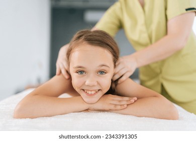 Closeup portrait of cheerful little girl having neck, shoulder and back massage by unrecognizable female pediatric masseuse at medical clinic, lying on massage table, looking at camera. - Powered by Shutterstock