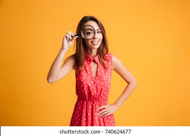 Close-up Portrait Of Cheerful Pretty Woman In Red Dress Looking At Camera Through Magnifying Glass, Isolated Over Yellow Background