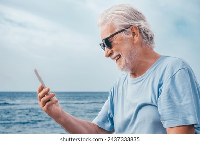 Close-up portrait of cheerful senior bearded man using smartphone in video call sitting outdoors at sea beach enjoying freedom, vacation or retirement concept - Powered by Shutterstock