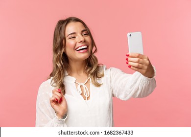 Close-up portrait of cheerful upbeat smiling blond girl, wearing white dress, laughing as record video, calling friend on mobile application, taking photo, selfie on smartphone, pink background
