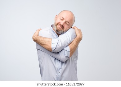 Closeup portrait of confident smiling man holding hugging himself isolated on grey wall background. Positive human emotion, facial expression. Love yourself concept