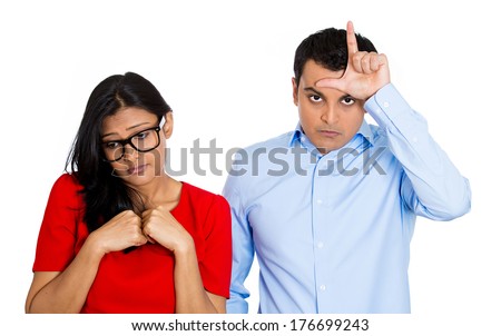 Closeup portrait of couple. Bully husband, man standing  upfront angry, loser sign on head, shy wife, nerdy woman wearing glasses looking downwards, isolated on white background. Human emotion culture