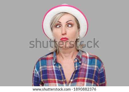 Closeup portrait of crazy crosseyed stylish mature woman in casual style with white hat standing, crossed eyes and looking with funny face expression. indoor studio shot isolated on gray background.