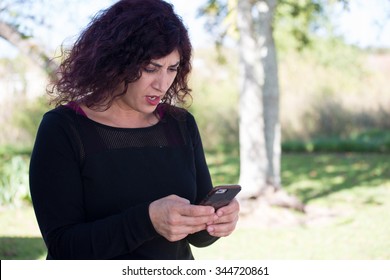 Closeup Portrait Curly Dark Hair Angry Young Woman Upset By What She Sees On Phone, Isolated Outside Green Trees Background. Negative Emotions Feelings